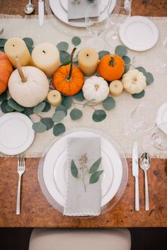 the table is set with white plates and silverware, pumpkins and greenery