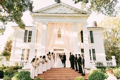 a group of people standing in front of a white house with a wedding party on the steps