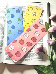 three colorful bookmarks sitting on top of an open book next to some pink flowers