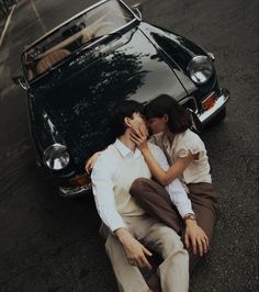 a man and woman sitting on the ground next to a black car with their arms around each other