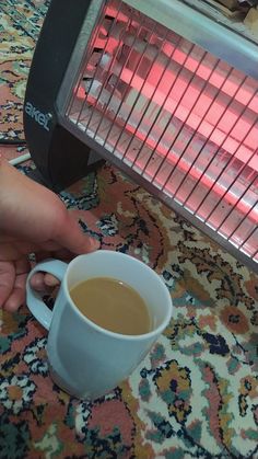 a person holding a cup of coffee in front of an electric heater on a rug