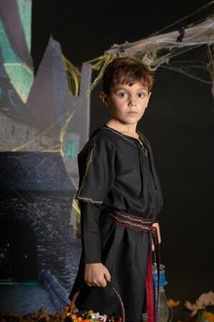 a young boy dressed in medieval clothing holding a basket and looking at the camera while standing next to a spider net