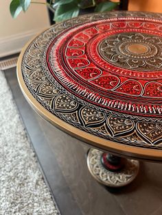 a red and gold decorated table on top of a carpeted floor next to a potted plant