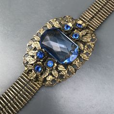 WIDE Vintage Art deco bracelet ~ with beautiful details with central large ,open-back faceted sapphire cab prong set on an gold tone openwork metal base ~ Embossed side link belt ~ Bracelet ends with hidden tongue and box clasp Dates 1930's - 1940 Measures : bracelet is 7 inches in wearable length x 1 3/4 inches max width . Condition : bracelet is in good old condition ~ Can see wear to gold gilding ,underneath silverish metal can be seen at places ~ Can see a small repair mark on the backside ( Wide Art, Rose Quartz Necklace Pendants, Art Deco Bracelet, Box Clasp, Message Jewelry, Gold Gilding, Flower Bracelet, Delicate Necklace, Vintage Art Deco