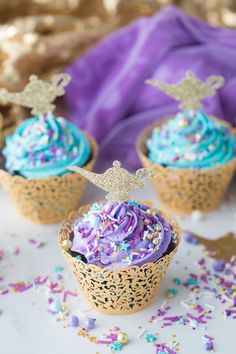 three cupcakes with blue frosting and purple sprinkles on a table