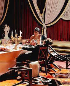 two people sitting at a table in front of red curtains and gold trimmings