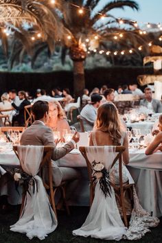 a group of people sitting at tables in front of palm trees with lights hanging from the ceiling