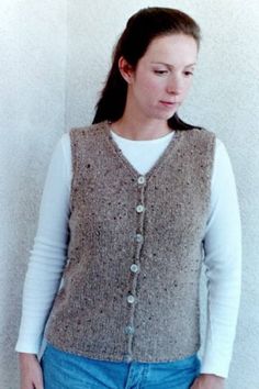 a woman standing in front of a white wall wearing a brown vest and blue jeans