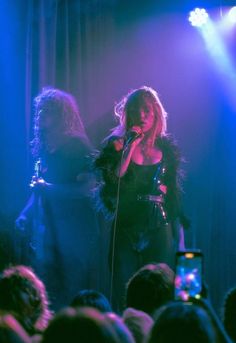 two women on stage singing into microphones with people in the back ground watching them