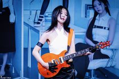 a woman holding an orange guitar in front of a wall with two women behind her