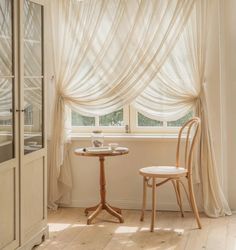 two chairs and a table in front of a window with sheer curtains on the windowsill