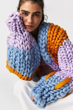 a woman sitting on a white surface with her hands behind her head, wearing a multicolored knitted scarf