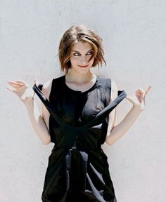 a woman with her hands in the air and wearing a black dress, standing against a white wall