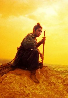 a man sitting on top of a dirt hill holding a stick and looking at the sky