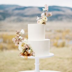 a three tiered white wedding cake with flowers on the top and bottom, sitting on a table outdoors