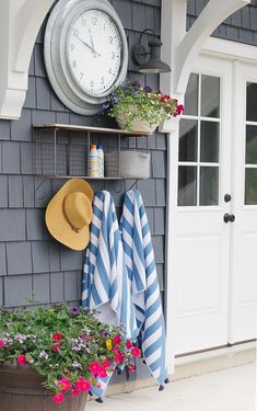 a clock mounted to the side of a gray house with flowers and towels hanging on it