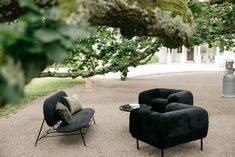 two black chairs sitting on top of a gravel covered ground next to a large tree