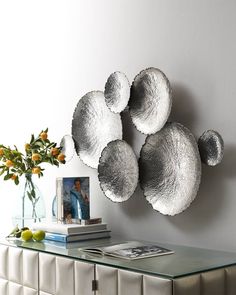 a glass table topped with silver plates and vase filled with oranges next to a white wall