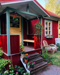 a small red house with green trim and flowers on the front porch, next to some chairs