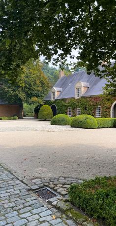 a large house with hedges in front of it on a cobblestone driveway surrounded by trees