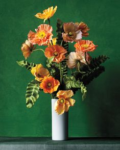 a white vase filled with lots of flowers on top of a green table next to a wall