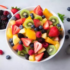 two bowls filled with fresh fruit on top of a white table next to berries and kiwis