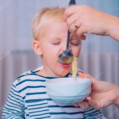 a little boy is eating from a bowl with his mother's hands and spoon