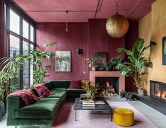 This living room is a bold study in color contrasts and texture. The rich, burgundy walls paired with the deep green velvet sofa create an opulent yet inviting atmosphere. Warmth radiates from the fireplace, while the lush plants and gold accents bring nature and luxury into perfect harmony. Every detail, from the oversized pendant to the pops of mustard and pink, makes this space feel both eclectic and elegant. It’s a true celebration of daring design, where each piece stands out yet fits se... Accent Wall For Dark Room, Dark Green Velvet Sofa Living Room Ideas, Bright Bold Living Room, Pink And Green Decor Living Room, Burgundy And Blush Living Room, Burgundy And Gold Living Room Ideas, Green Burgundy Living Room, Green And Maroon Living Room, Contrast Fireplace