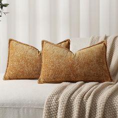 two gold pillows sitting on top of a white couch next to a potted plant