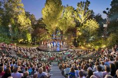 an outdoor theater with people sitting on the stage and in front of it is lit up at night