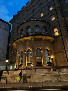 a man walking down the street in front of a large building at night with lights on