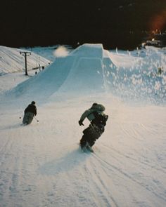 two snowboarders going down a snowy hill at night with the sun shining on them