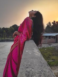 a woman in a pink sari leaning on a concrete wall looking up at the sky