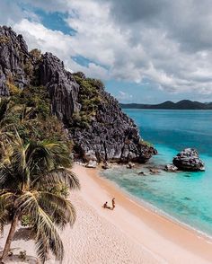 two people are walking on the beach next to some rocks and palm trees in the water