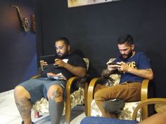 two men sitting in chairs with their legs crossed and one is looking at his cell phone