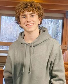 a young man with curly hair standing in front of a window wearing a gray hoodie