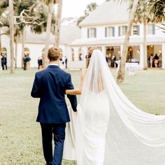 a bride and groom walking through the grass with their wedding veil blowing in the wind