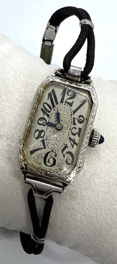 "This scarce lady's Art Deco watch features the coveted dial of exploding numbers.  By Belais, the case is 18K white gold, completely engraved, and is in perfect condition.  I don't know how much of the design on the back of the case was custom, but someone went all out in monograming.  The crown is set with a sapphire glass cabochon.  The black cord band with an Art Deco design on the clasp may or may not be original--the clasp mechanism is stamped with a patent number that dates it to 1937.  If the watch itself dates earlier, then the band is a later addition.  Judging the band, case, and crystal, I would guess that the watch had never been worn.  The dial with the fabulous exploding numbers does show some oxidation at the bottom, which isn't as bad in person as it looks in the photos. Timeless Engraved Watches For Anniversary, Elegant Collectible Watch With Rectangular Dial, Luxury Engraved Collectible Jewelry And Watches, White Gold Automatic Watches For Anniversary, Antique Silver Watch With Rectangular Dial, Antique Silver Watches With Rectangular Dial, Engraved Watch With Rectangular Dial For Anniversary, Engraved Watches With Rectangular Dial For Anniversary, Collectible Timeless White Gold Watches