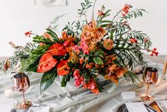 an arrangement of flowers and greenery is displayed on a white table cloth with silverware