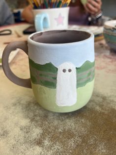 a coffee cup with a ghost painted on it sitting on a table next to some paintbrushes