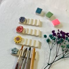 several different types of art supplies and flowers on a white tablecloth with watercolor paints