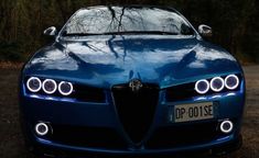 the front end of a blue car with its lights turned on and trees in the background