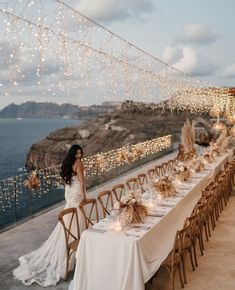 a woman standing next to a long table covered in white cloths with lights strung over it