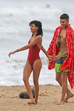 a man and woman walking on the beach with towels over their shoulders, holding hands