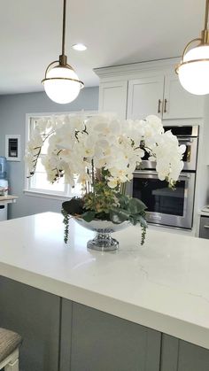 white flowers in a glass bowl on a kitchen countertop with lights above it and an oven