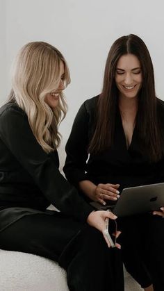 two women sitting on a white couch looking at a tablet computer screen and laughing together