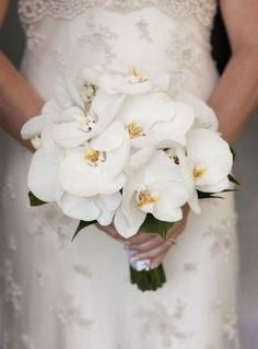 a woman in a white dress holding a bouquet of white orchids and greenery