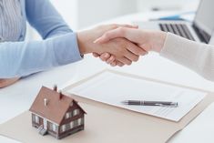 two people shaking hands in front of a house on a desk with a pen and paper