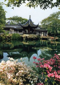 a pond surrounded by trees and flowers with a building in the background that has a pagoda on top