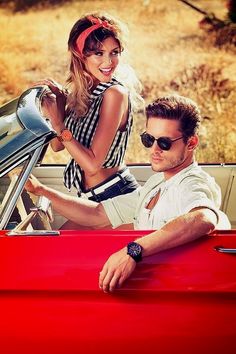 a man and woman sitting in the back of a red convertible car, posing for a photo